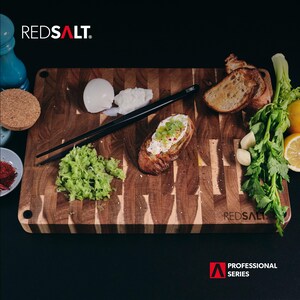 May include: A wooden cutting board with a checkered pattern, featuring a piece of toasted bread with cream cheese and green toppings, a small pile of chopped green vegetables, and a pair of black chopsticks. The board is labeled "REDSALT. PROFESSIONAL SERIES".