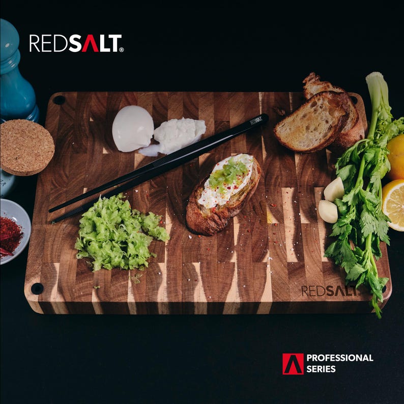May include: A wooden cutting board with a checkered pattern, featuring a piece of toasted bread with cream cheese and green toppings, a small pile of chopped green vegetables, and a pair of black chopsticks. The board is labeled "REDSALT. PROFESSIONAL SERIES".