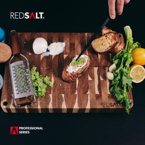 May include: A wooden cutting board with a variety of ingredients for preparing a meal, including a grater, bread, cheese, and herbs. The board is labeled "REDSALT" and features the text "PROFESSIONAL SERIES".