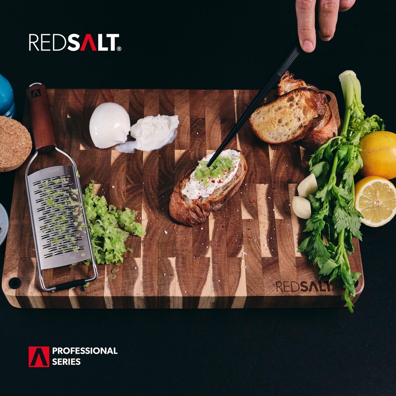May include: A wooden cutting board with a variety of ingredients for preparing a meal, including a grater, bread, cheese, and herbs. The board is labeled "REDSALT" and features the text "PROFESSIONAL SERIES".