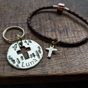 May include: A brown leather bracelet with a gold cross charm and a silver key chain with a round charm featuring a cross, trees, and the name "Luna".