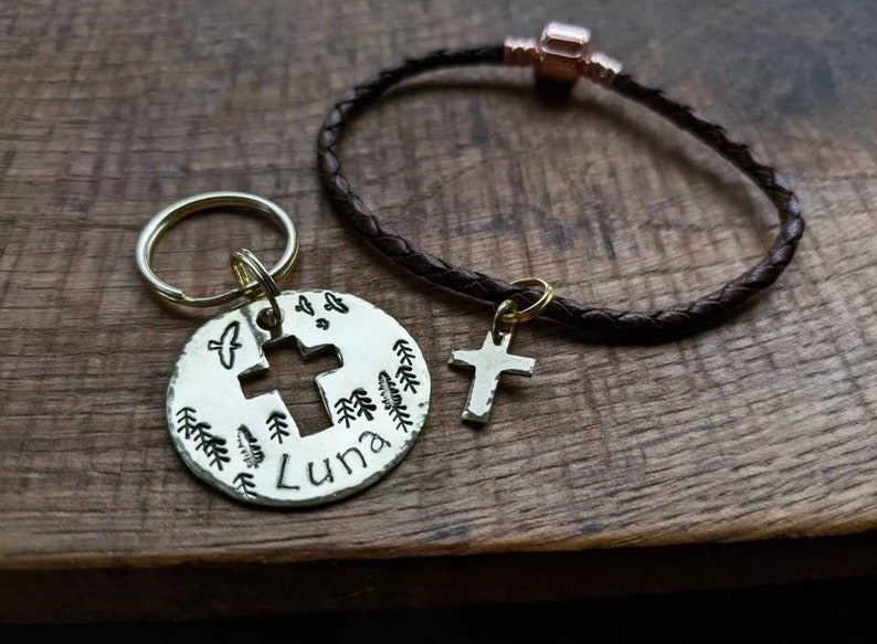 May include: A brown leather bracelet with a gold cross charm and a silver key chain with a round charm featuring a cross, trees, and the name "Luna".