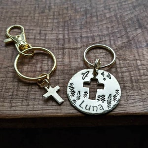 May include: A silver key chain with a cross cut-out and the name "Luna" engraved on a round tag. The tag is surrounded by small tree designs. The key chain is attached to a gold split ring.