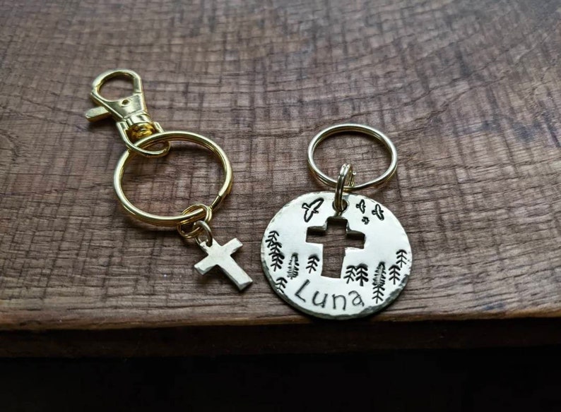 May include: A silver key chain with a cross cut-out and the name "Luna" engraved on a round tag. The tag is surrounded by small tree designs. The key chain is attached to a gold split ring.
