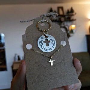 May include: Gold necklace with a small cross charm and a larger round charm with a cross and the name "Harlee" engraved on it. The charms are attached to a gold chain and a key ring.