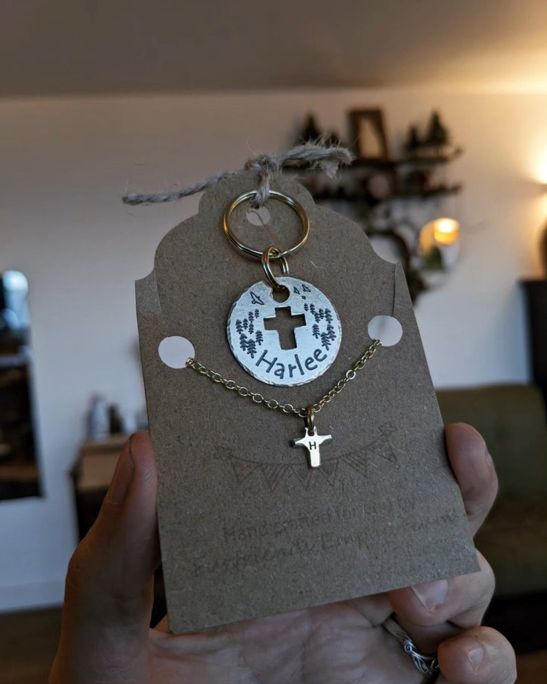 May include: Gold necklace with a small cross charm and a larger round charm with a cross and the name "Harlee" engraved on it. The charms are attached to a gold chain and a key ring.
