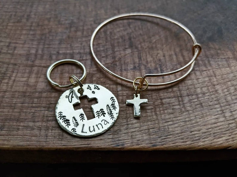 May include: A gold charm bracelet with a cross charm and a gold key chain with a round charm featuring a cross, trees, and the name "Luna".