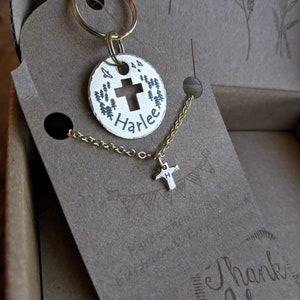 May include: A gold-toned keychain with a round charm featuring a cross and the name "Harlee" engraved on it. The charm is attached to a gold-toned key ring. A matching gold-toned necklace with a small cross charm is also included.