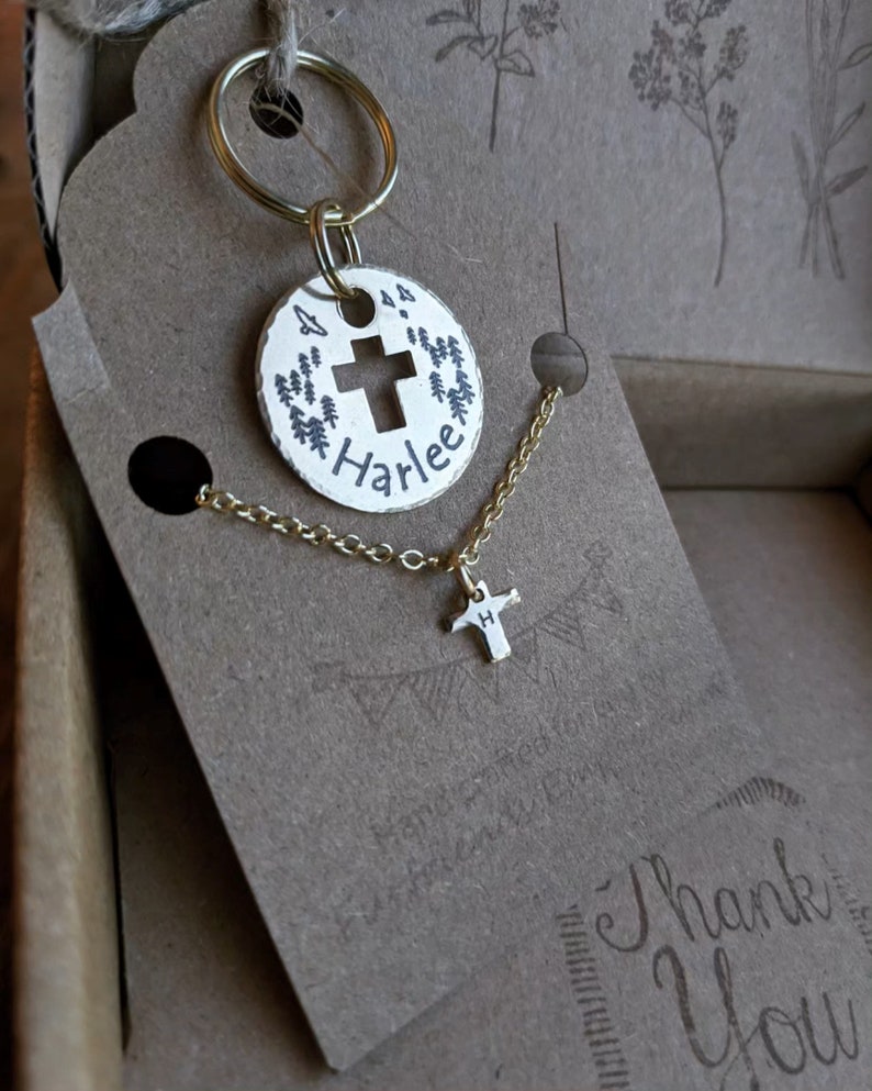 May include: A gold-toned keychain with a round charm featuring a cross and the name "Harlee" engraved on it. The charm is attached to a gold-toned key ring. A matching gold-toned necklace with a small cross charm is also included.