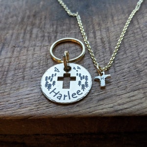 May include: A silver charm with a cross and the name "Harlee" engraved on it, surrounded by small pine trees. The charm is attached to a gold key ring. A separate gold chain with a small cross charm is also pictured.