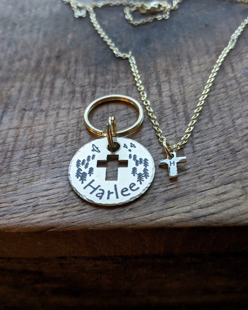 May include: A silver charm with a cross and the name "Harlee" engraved on it, surrounded by small pine trees. The charm is attached to a gold key ring. A separate gold chain with a small cross charm is also pictured.