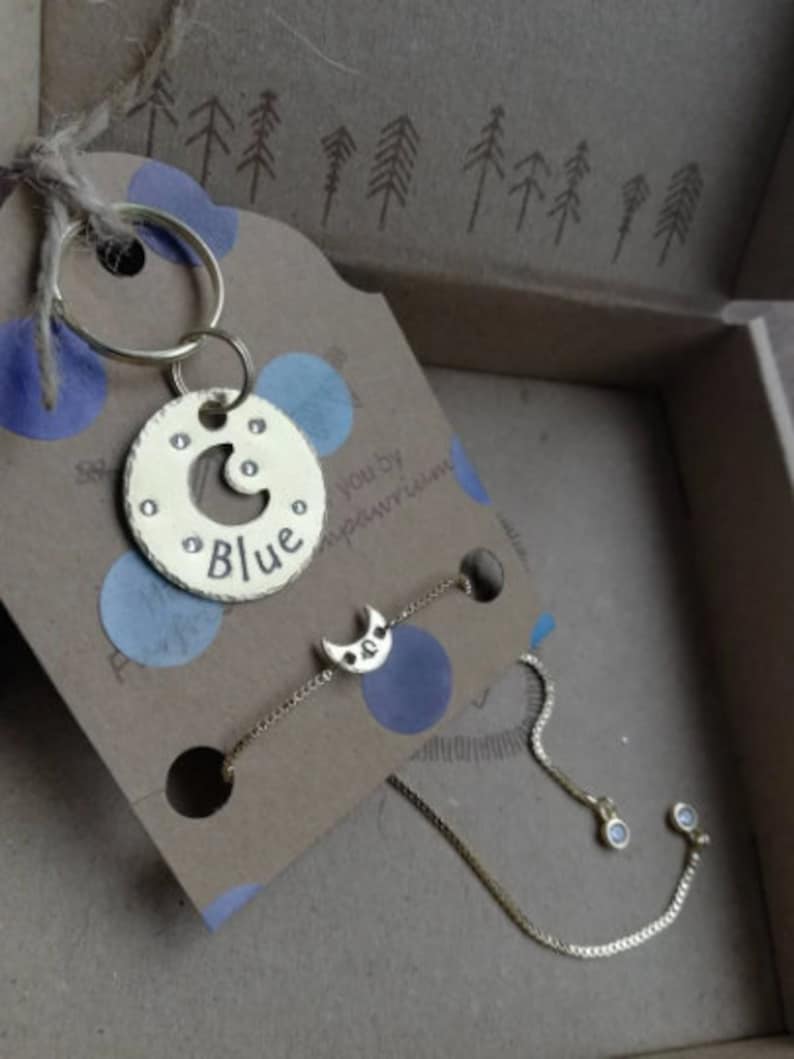 Puede incluir: Un llavero dorado con un dije de media luna y la palabra "Blue" grabada en una etiqueta redonda. El llavero est&aacute; empaquetado en una caja con papel estampado azul y blanco.