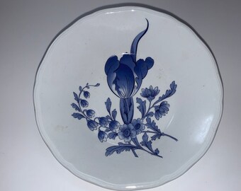 May include: A white ceramic saucer with a blue floral design. The design features a large blue flower with a stem and leaves, surrounded by smaller blue flowers.