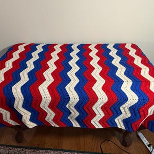 May include: A handmade crochet blanket with a bold chevron pattern in red, white, and blue. The blanket is draped over a wooden table, showcasing its vibrant colors and textured design. This cozy blanket is perfect for adding a touch of handmade charm to any home.