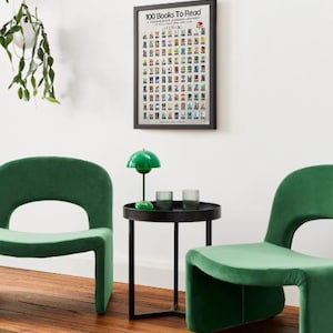 May include: A framed poster titled "100 Books To Read" hangs on a white wall. Below, two green velvet chairs flank a small black table with a green lamp and two glasses. A hanging plant is visible on the left.