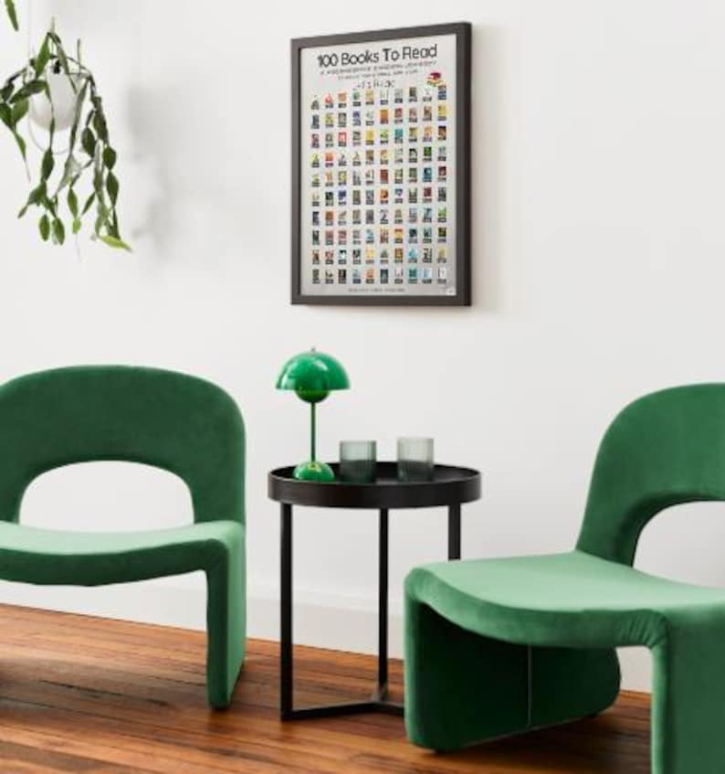 May include: A framed poster titled "100 Books To Read" hangs on a white wall. Below, two green velvet chairs flank a small black table with a green lamp and two glasses. A hanging plant is visible on the left.