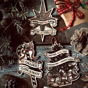 May include: Three wooden Christmas ornaments with dark, hand-drawn illustrations and red accents. The first ornament is a star with the text "RIP Probably Dead 4000 Years Before You Gazed At-It". The second ornament is a candy cane with the text "60 Calories Closer To Death". The third ornament is a Christmas tree with the text "Another Dead Tree Contributing To Climate Change".