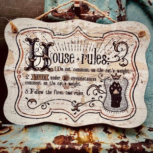 May include: A rustic wooden sign with the text "House-rules:" and three rules written in black text. The rules are: 1. Do not comment on the cat's weight. 2. NEVER under NO circumstances comment on the cat's weight. 3. Follow the first two rules.