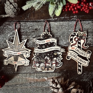 May include: Three wooden Christmas ornaments with humorous sayings. The first ornament is a star with the text "RIP Probably Dead 4000 Years Before You Gazed At It". The second ornament is a Christmas tree with the text "Another Dead Tree Contributing To Climate Change". The third ornament is a candy cane with the text "60 Calories Closer To Death".