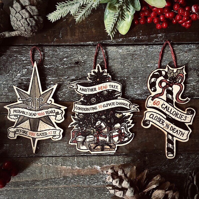 May include: Three wooden Christmas ornaments with humorous sayings. The first ornament is a star with the text "RIP Probably Dead 4000 Years Before You Gazed At It". The second ornament is a Christmas tree with the text "Another Dead Tree Contributing To Climate Change". The third ornament is a candy cane with the text "60 Calories Closer To Death".