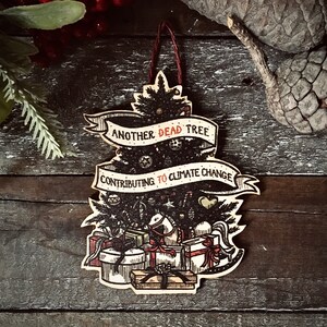 May include: A wooden Christmas tree ornament with a black and white illustration of a Christmas tree with the text "Another Dead Tree Contributing to Climate Change".