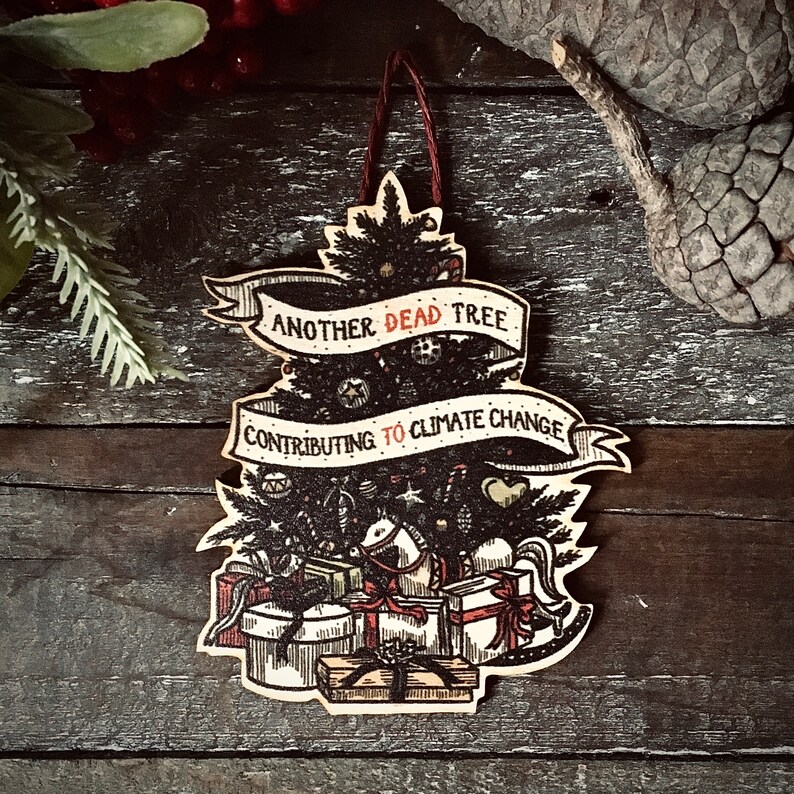 May include: A wooden Christmas tree ornament with a black and white illustration of a Christmas tree with the text "Another Dead Tree Contributing to Climate Change".
