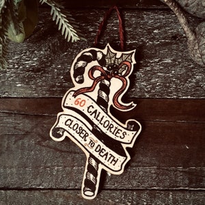May include: A wooden Christmas ornament shaped like a candy cane with a bat on top. The ornament is decorated with black and white stripes and red ribbon. The text on the ornament reads "60 Calories Closer to Death".