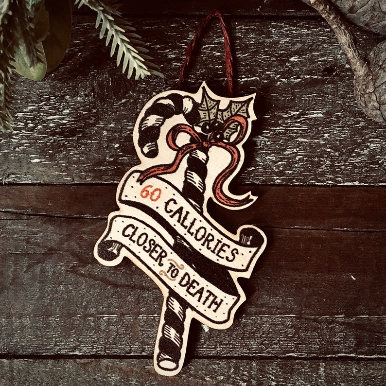 May include: A wooden Christmas ornament shaped like a candy cane with a bat on top. The ornament is decorated with black and white stripes and red ribbon. The text on the ornament reads "60 Calories Closer to Death".