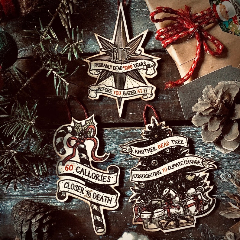 May include: Three wooden Christmas ornaments with dark, hand-drawn illustrations and red accents. The first ornament is a star with the text "RIP Probably Dead 4000 Years Before You Gazed At-It". The second ornament is a candy cane with the text "60 Calories Closer To Death". The third ornament is a Christmas tree with the text "Another Dead Tree Contributing To Climate Change".