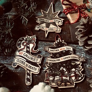 May include: Four handmade Christmas ornaments with dark brown wood backgrounds and black ink illustrations. The ornaments feature the text "RIP Probably Dead 4000 Years Before You Gazed At It", "60 Calories Closer To Death", "Another Dead Tree Contributing To Climate Change", and a candy cane.