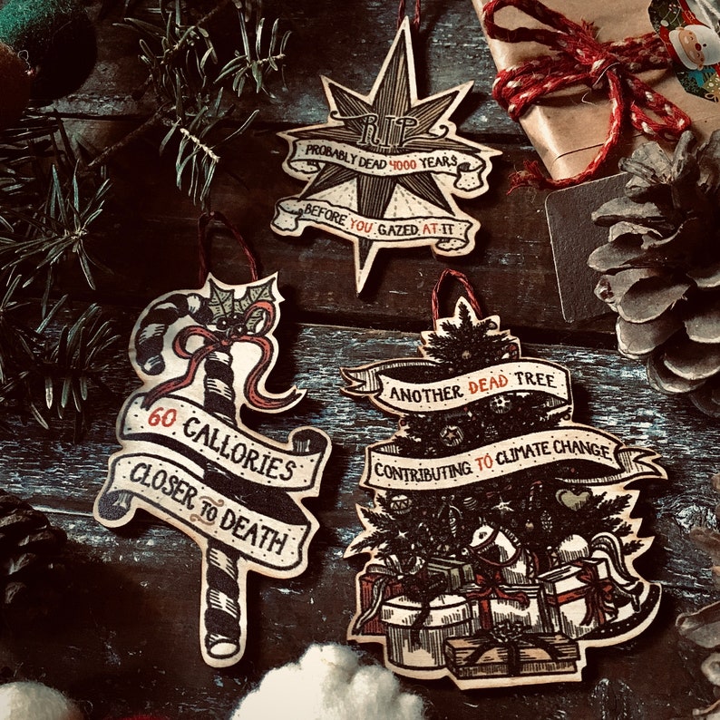 May include: Four handmade Christmas ornaments with dark brown wood backgrounds and black ink illustrations. The ornaments feature the text "RIP Probably Dead 4000 Years Before You Gazed At It", "60 Calories Closer To Death", "Another Dead Tree Contributing To Climate Change", and a candy cane.