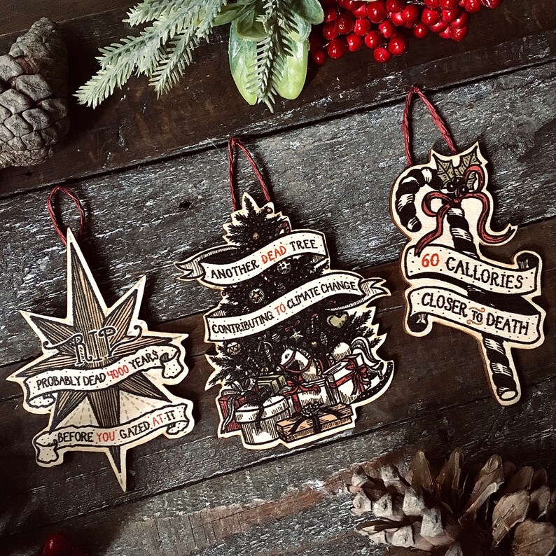 May include: Three Christmas ornaments with humorous sayings. The first ornament is a star with the text "Probably dead 4000 years before you gazed at it". The second ornament is a Christmas tree with the text "Another dead tree contributing to climate change". The third ornament is a candy cane with the text "60 Calories closer to death".