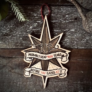 May include: A wooden star-shaped ornament with a black and white illustration of a skull and crossbones. The ornament has a red ribbon and the text "RIP Probably Dead 4000 Years Before You Gazed At It".