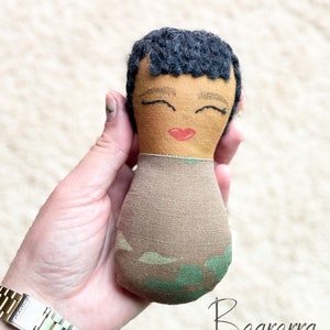May include: A handmade fabric doll with a tan face, black hair, and closed eyes. The doll has a red painted mouth and is wearing a brown and green patterned outfit. The doll is held in a hand.