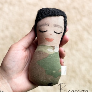 May include: A handmade doll with black hair, closed eyes, and a pink mouth. The doll is wearing a camouflage fabric outfit.