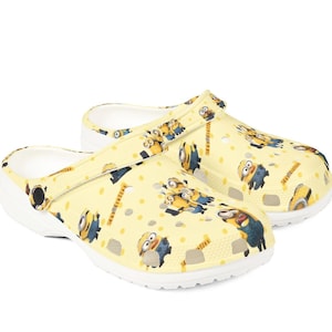May include: A pair of yellow clogs featuring a repeating pattern of Minion characters. The clogs have a white sole and a strap that can be worn around the heel or across the top of the foot. The background is white.