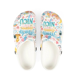 May include: White clogs with a colorful pattern featuring the text "NICU" and "Miracles Happen in the NICU." The design includes hearts, rainbows, a giraffe, and medical equipment. The clogs have a white sole and black accents.