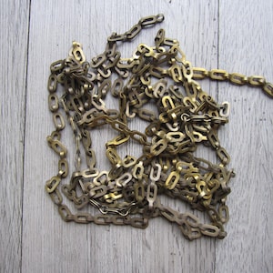 May include: A tangled pile of antique brass-colored chain. The chain links are oval-shaped and vary slightly in color, from a darker bronze to a brighter gold. The chain is lying on a light gray wooden surface.