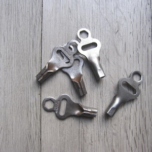 May include: A collection of vintage metal keys with a unique design, featuring a hexagonal loop and a slotted opening. The keys are arranged on a light gray wooden surface, showcasing their aged, metallic appearance.