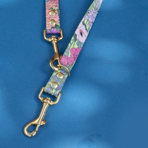 May include: A floral dog lead with gold-tone metal clasps and rivets. The lead has a colourful floral pattern in pink, purple, and orange on a green background. The lead is coiled and displayed on a blue surface.