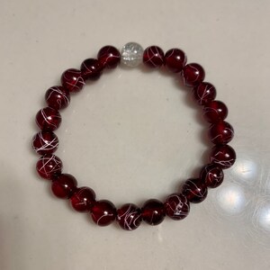 May include: A red beaded bracelet with white swirls on each bead. The bracelet has a clear crystal bead in the center.