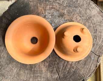 May include: Two terracotta clay pots, one with a smooth, rounded bottom and the other with three small feet. Both pots have a drainage hole in the center.