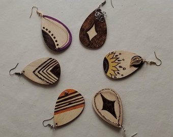 May include: Five pairs of wooden earrings with different geometric and abstract designs. The earrings are all teardrop shaped and have a natural wood finish. Some of the earrings have a gold accent.