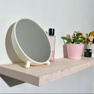 May include: A white shelf with a round white mirror, a pink glass bottle, a pink flower pot with green leaves, and a white container with makeup brushes.