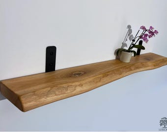 Puede incluir: Una estantería de madera natural con un borde vivo y un acabado rugoso y rústico. La estantería está montada en la pared con soportes de metal negro. Una pequeña planta en maceta con flores rosas se encuentra sobre la estantería.
