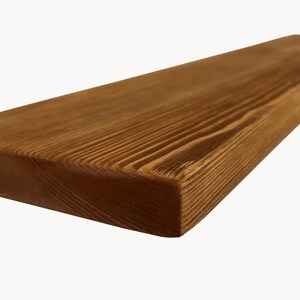 May include: Close-up of a rectangular wooden shelf with a natural wood grain. The shelf is a warm brown color and has a smooth, finished surface. The edges are slightly rounded. This shelf is suitable for home decor.