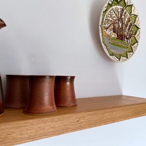 Pu&ograve; includere: Uno scaffale in legno con una brocca marrone e tre tazze marroni in cima. Lo scaffale &egrave; montato su una parete bianca. Un piatto decorativo con un design verde e marrone &egrave; appeso al muro sopra lo scaffale.