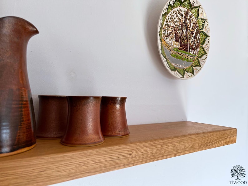 Pu&ograve; includere: Uno scaffale in legno con una brocca marrone e tre tazze marroni in cima. Lo scaffale &egrave; montato su una parete bianca. Un piatto decorativo con un design verde e marrone &egrave; appeso al muro sopra lo scaffale.