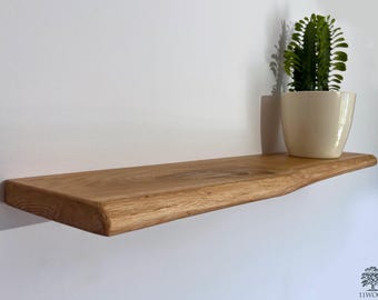 May include: A natural wood shelf with a live edge, stained in a light brown color. The shelf is mounted to a white wall and has a plant in a white pot on top of it.