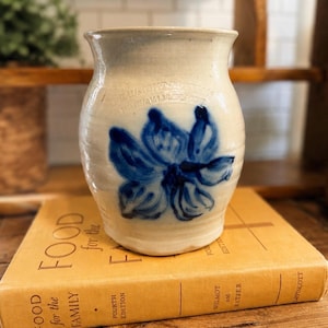 May include: A beige stoneware crock featuring a hand-painted blue floral design on the front. The vessel has a slightly flared rim and a rounded body. Faint, embossed text is visible near the top rim, and the crock sits on a yellow book.